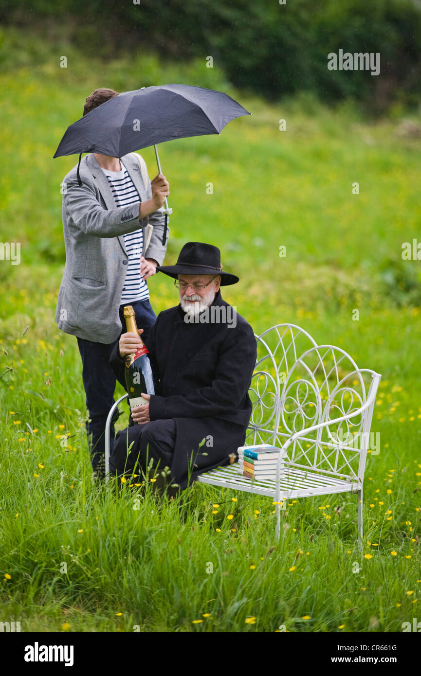 Terry Pratchett being sheltered from rain with umbrella on photo shoot at The Telegraph Hay Festival 2012, Hay-on-Wye, Powys, Wales, UK Stock Photo