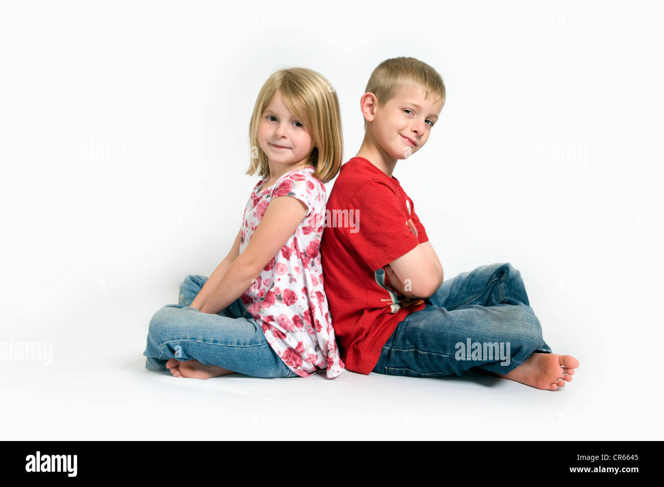 Two Caucasian children, a brother and sister sat back to back smiling (7 year old girl and 8 year old boy) on a white background Stock Photo