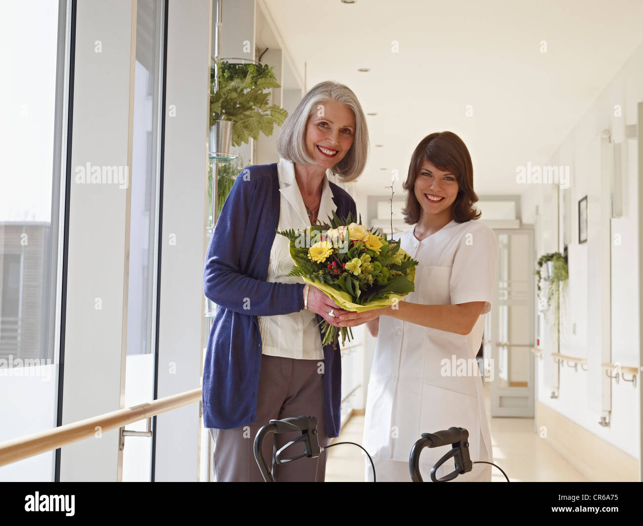 Germany, Cologne, Senior women and caretaker holding bouquet in corridor of nursing room, smiling, portrait Stock Photo