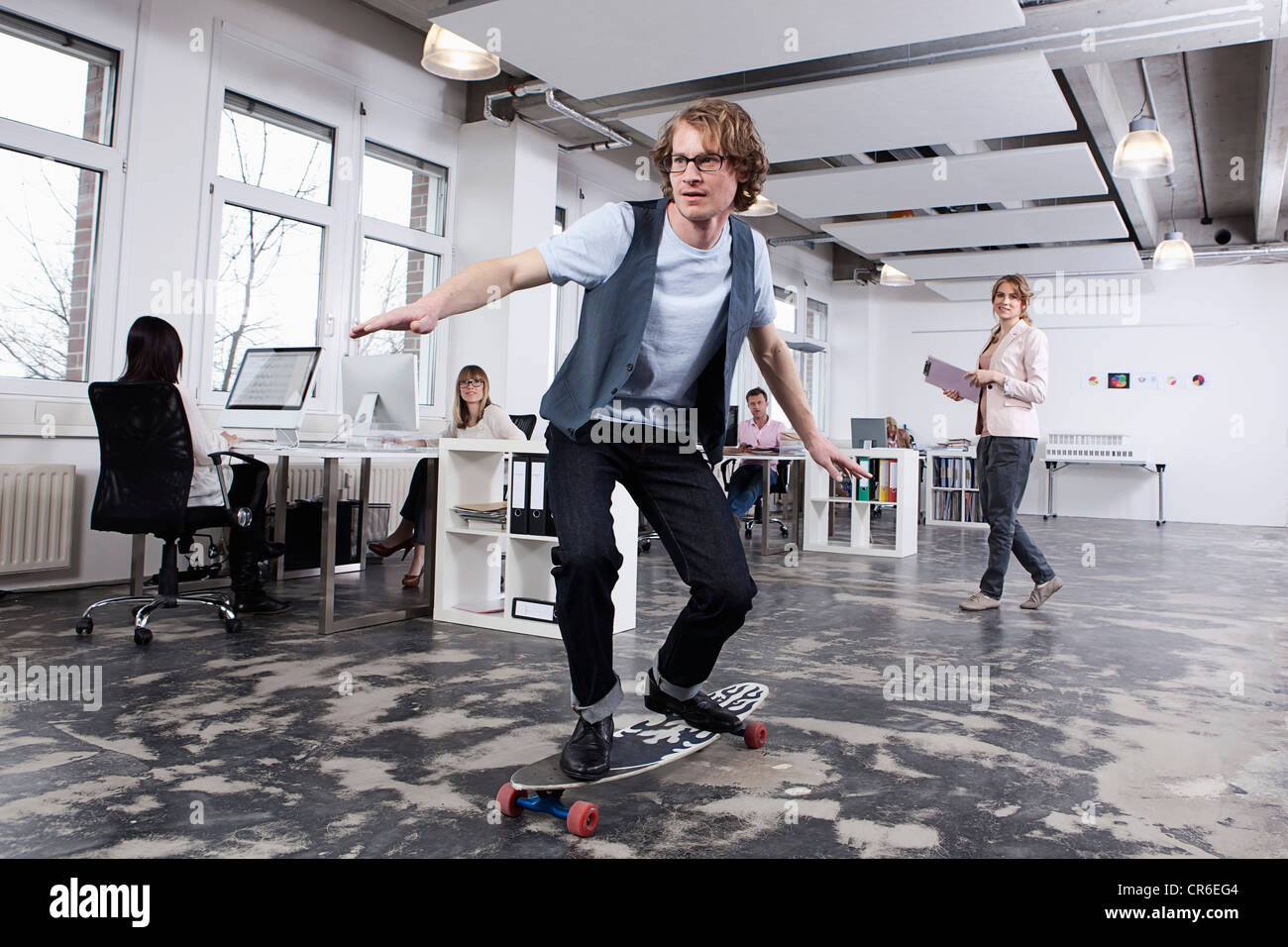 Germany, Bavaria, Munich, Man skate boarding in office while colleagues working in background Stock Photo
