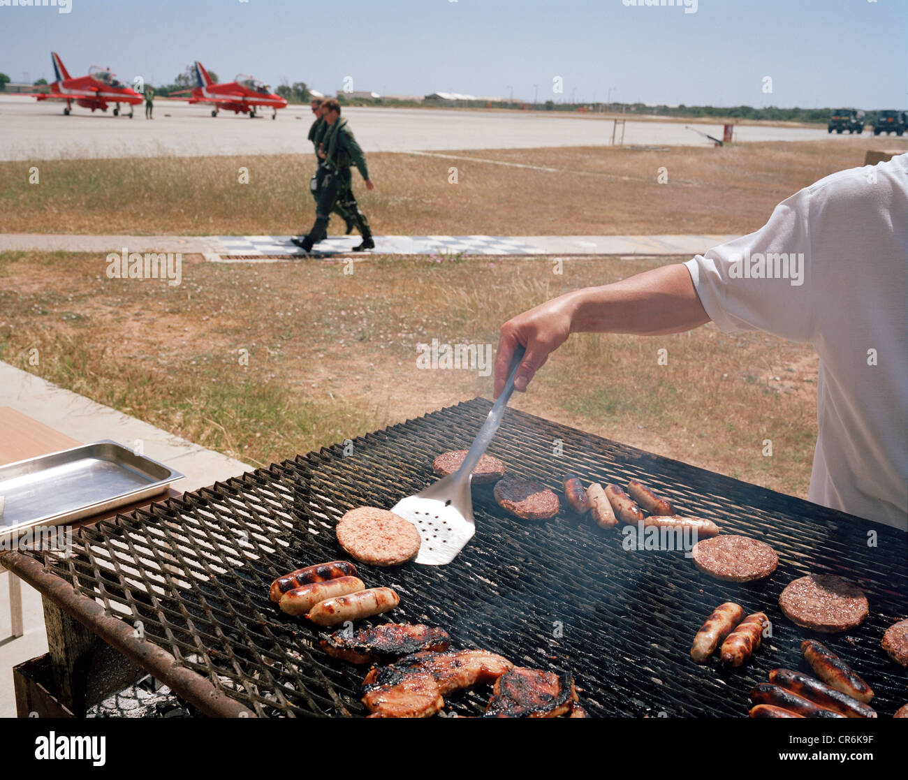 Anonymous chef prepares BBQ burgers and sausages as a pilot of the Red Arrows, Britain's RAF aerobatic team walks past. Stock Photo