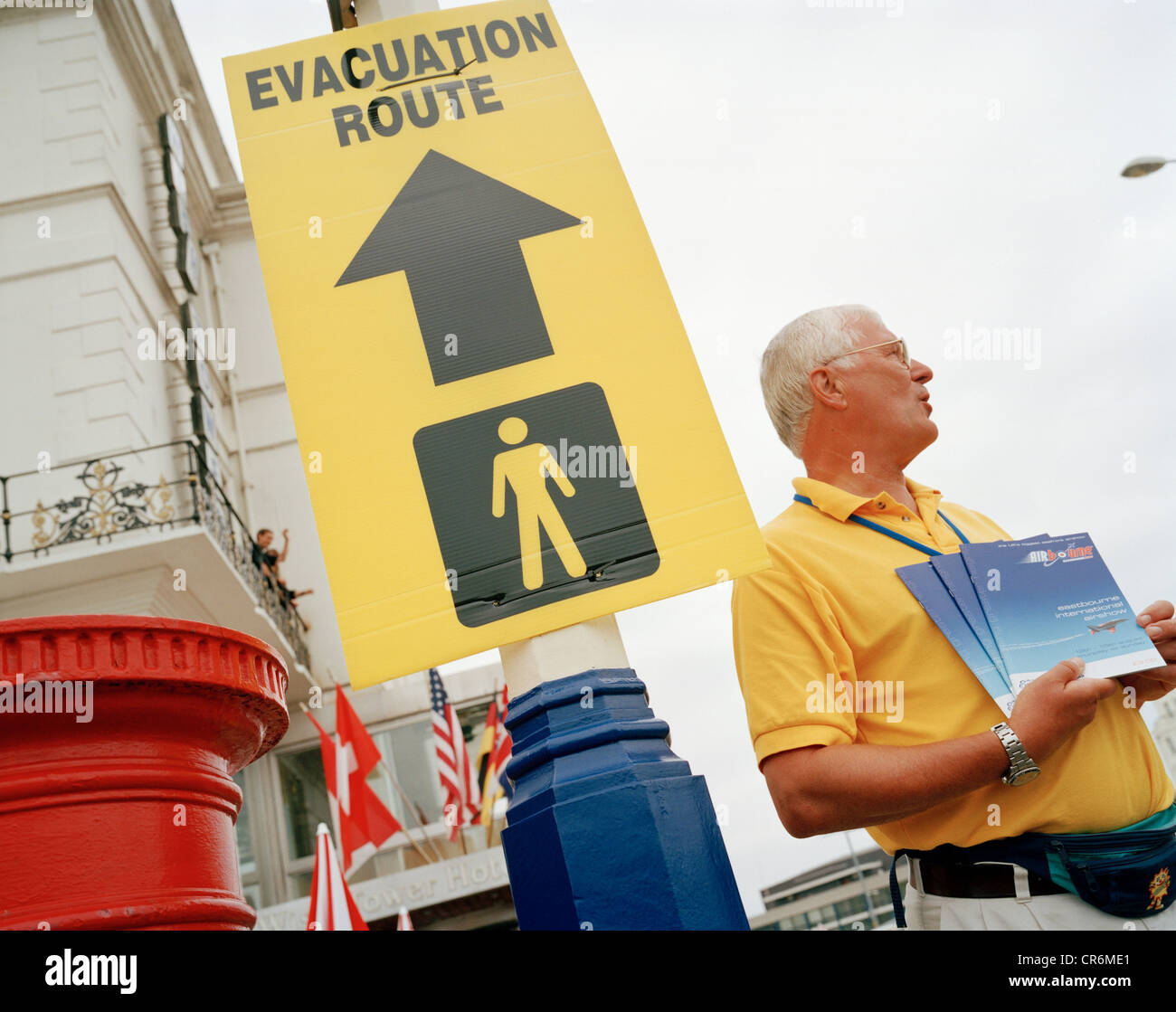 Man sells airshow programmes under council sign for evacuation routes poster during local airshow weekend. Stock Photo