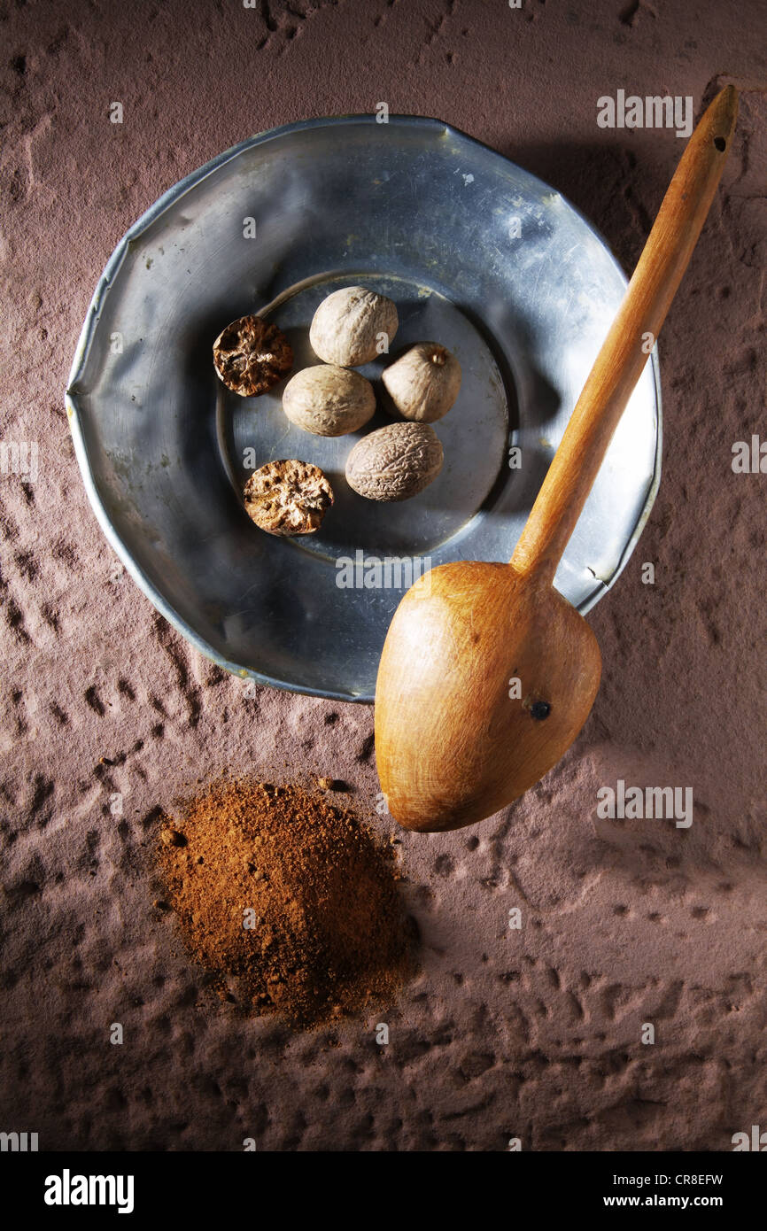 Nutmegs (Myristica fragrans), on a metal plate with a wooden spoon and ground nutmeg on a rustic stone base Stock Photo