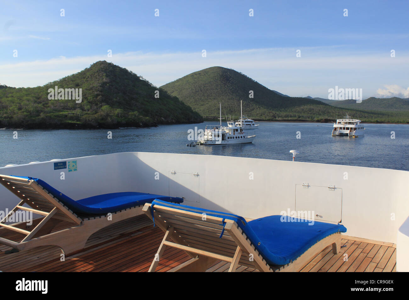 Two empty sun loungers on deck of small boat overlooking Floreana island and three ships Galapagos Islands Ecuador Stock Photo