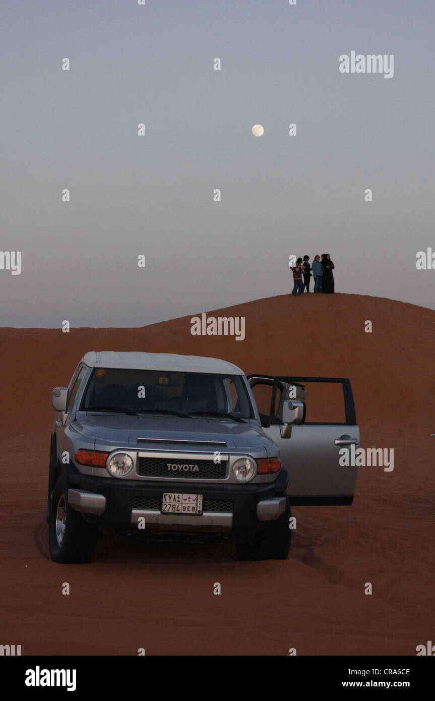 The moon over sand dunes and a group of people standing on the top of dune, a four wheel drive vehicle in foreground, Riyadh KSA Stock Photo
