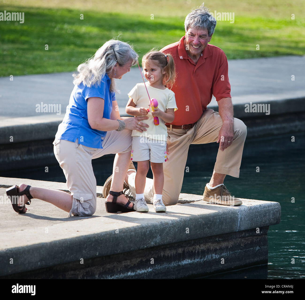 Grandmother and grandfather play with granddaughter at park by local pond. Stock Photo