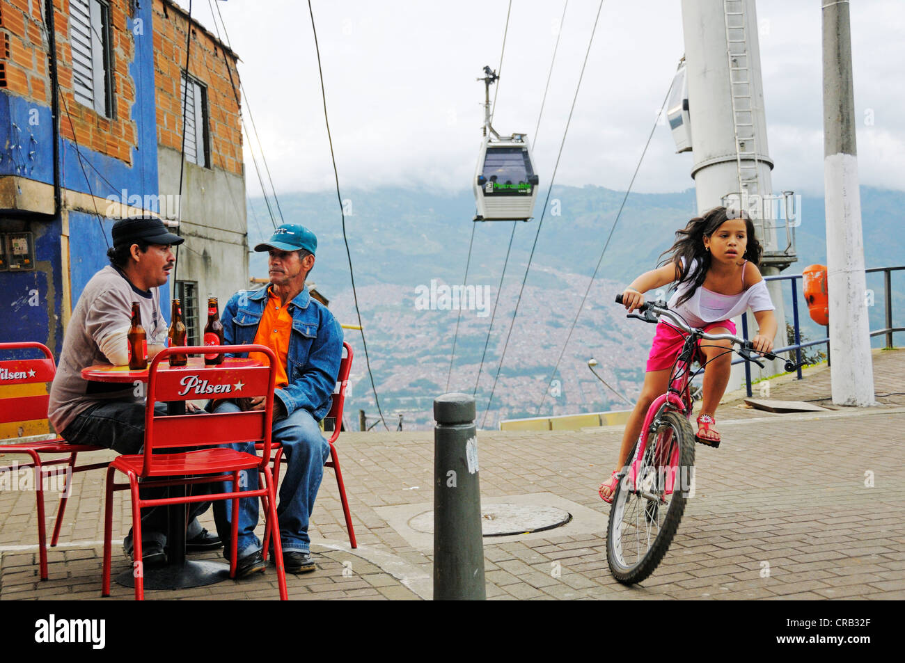 Men sitting in a street café, girl riding a bicycle, Metrocable cable car at back, slums, Comuna 13, Medellin, Colombia Stock Photo