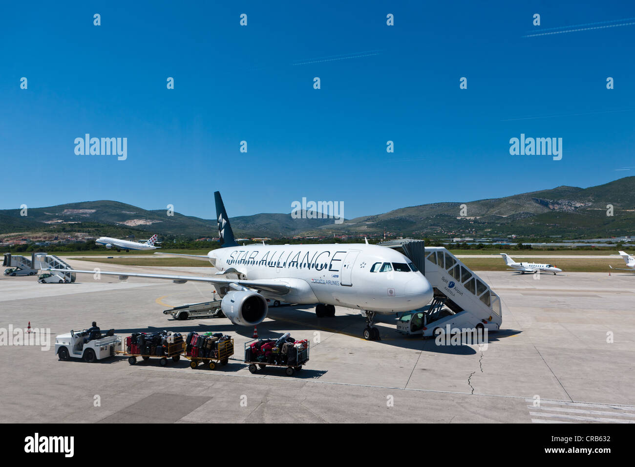 Star Alliance Airbus A319 aircraft at Split Airport, with a luggage cart at the front and a plane taking-off at the rear, Split Stock Photo