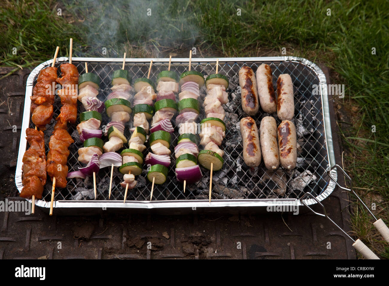Disposable charcoal barbecue cooking kebabs and sausages. Stock Photo