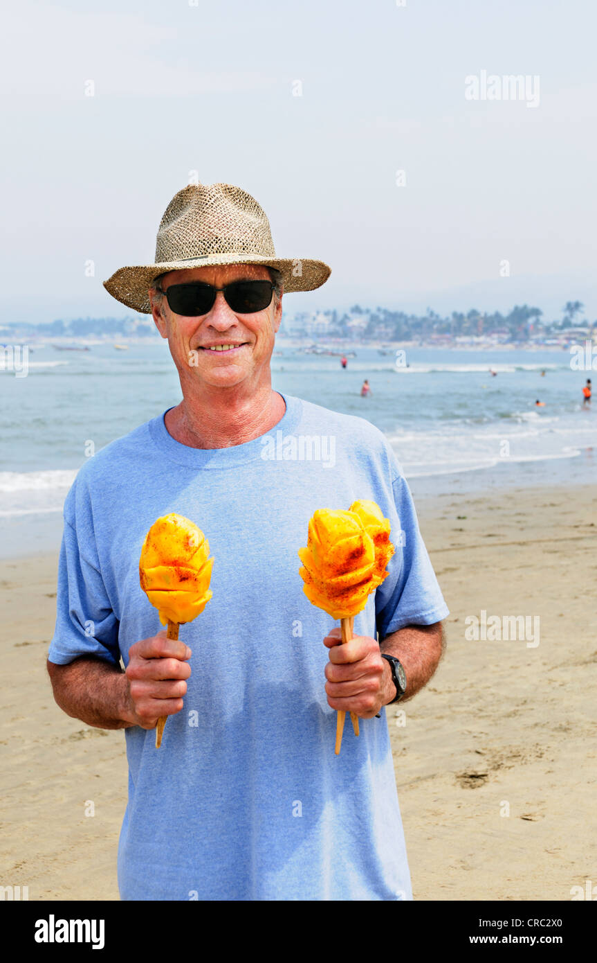 Adult senior male holds two mangoes on a stick on the beach in Guayabitos, Nayarit,  Mexico. Stock Photo