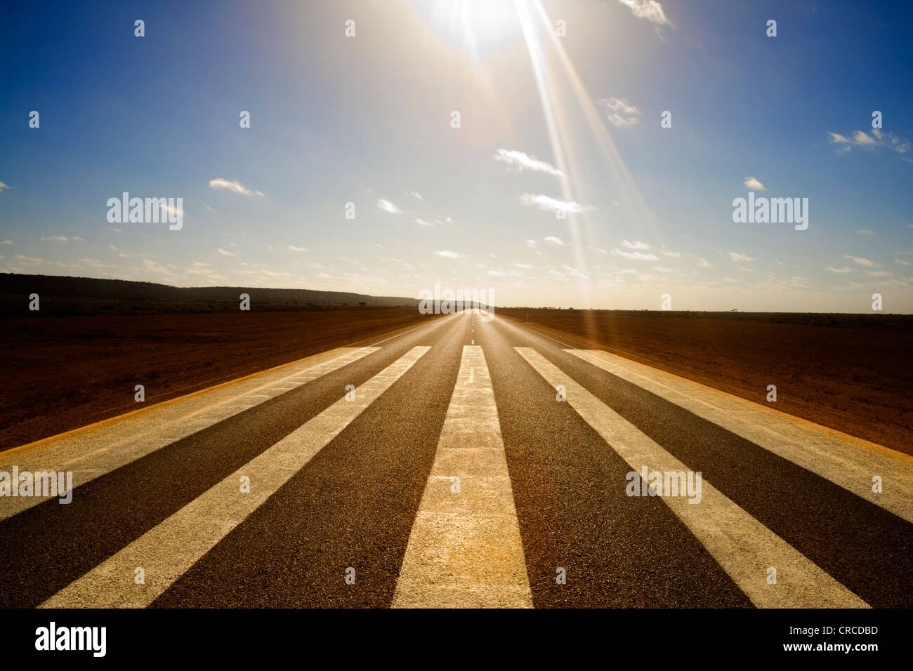 Wide angle shot of long straight road with runway markings and sun in picture causing lens flare. Stock Photo