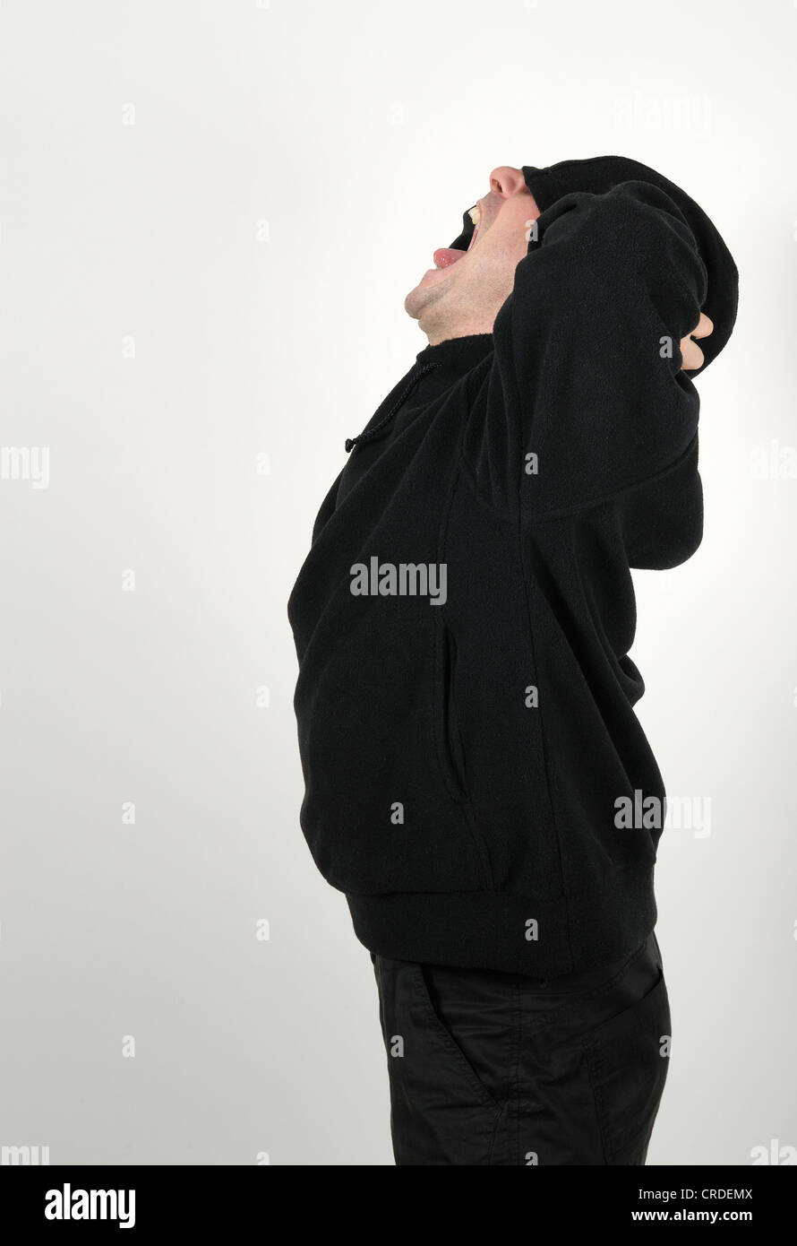 Young man wearing a black hooded shirt with his eyes under the hood, screaming madly and desperately Stock Photo
