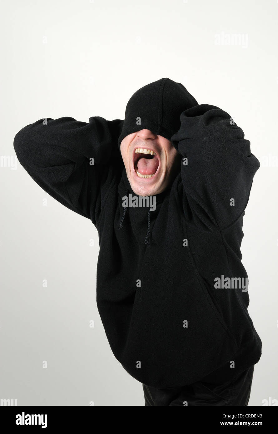 Young man wearing a black hooded shirt with his eyes under the hood, screaming madly and desperately Stock Photo