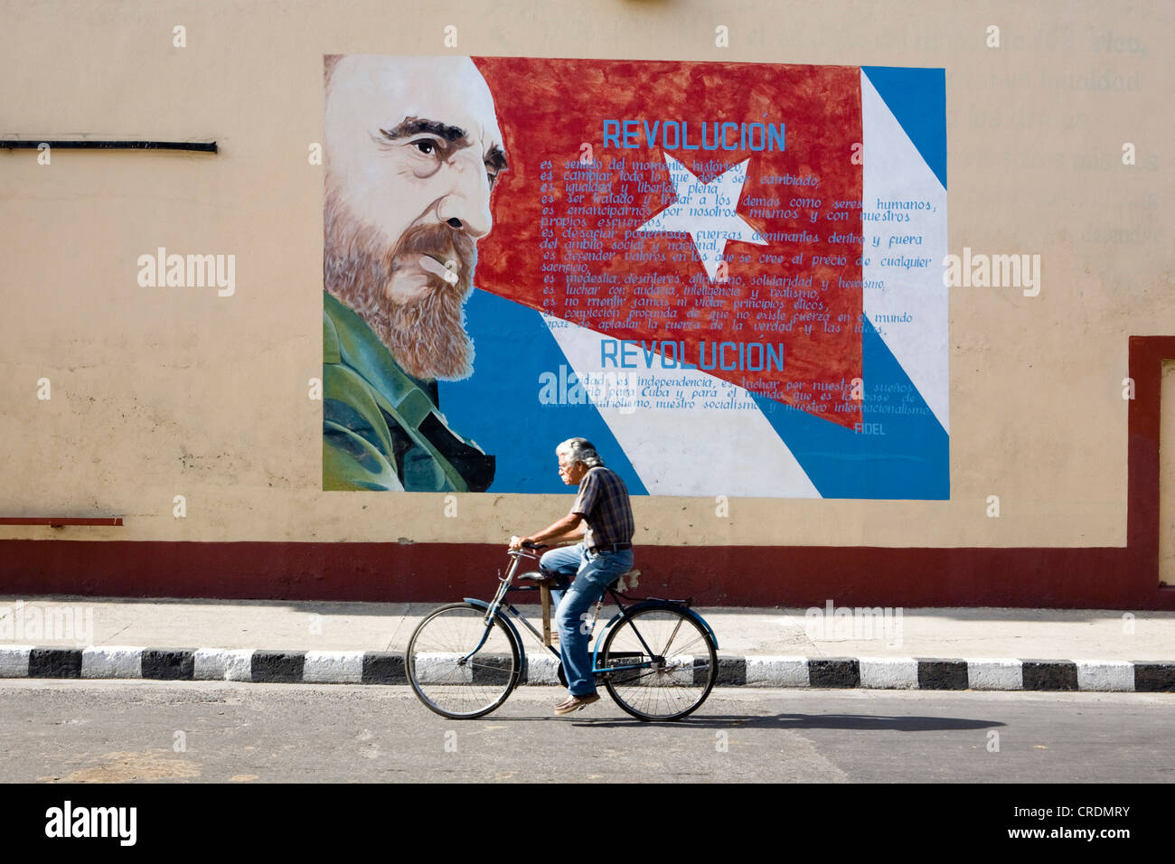 Old cyclist cycling in front of revolutionary propaganda on a facade with picture of Fidel Castro, Cienfuegos, Cuba Stock Photo