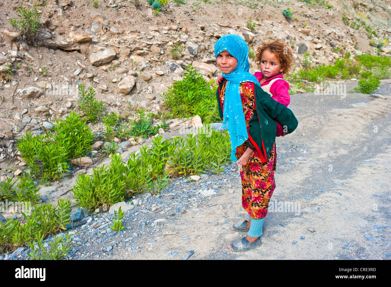 Young girl wearing a blue head scarf and carrying her younger sister in a sling on her back, High Atlas Mountains, Morocco Stock Photo