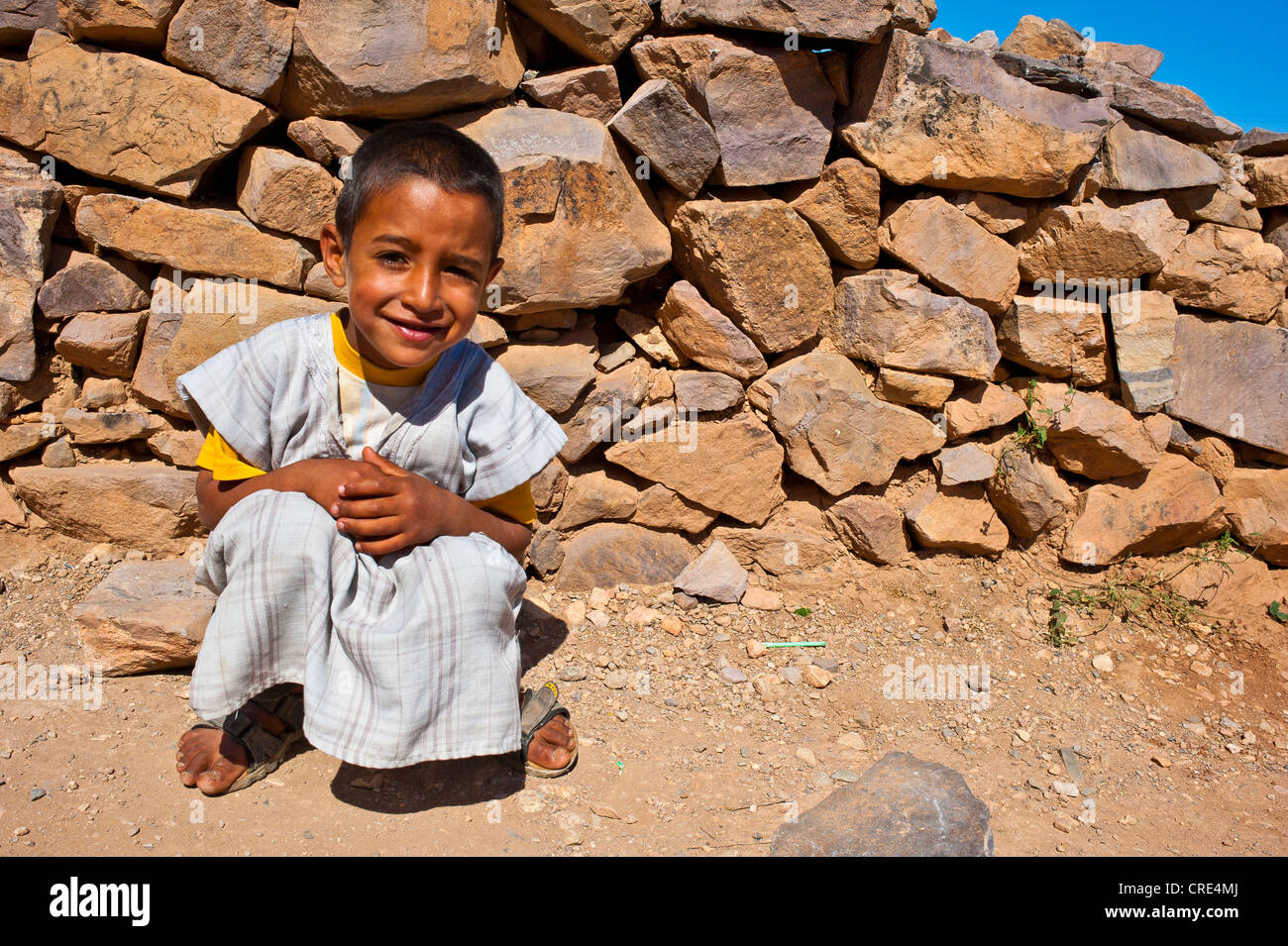 Small friendly boy wearing a traditional djellabah garment squatting in front of a stone wall, Agadir Tasguent Stock Photo