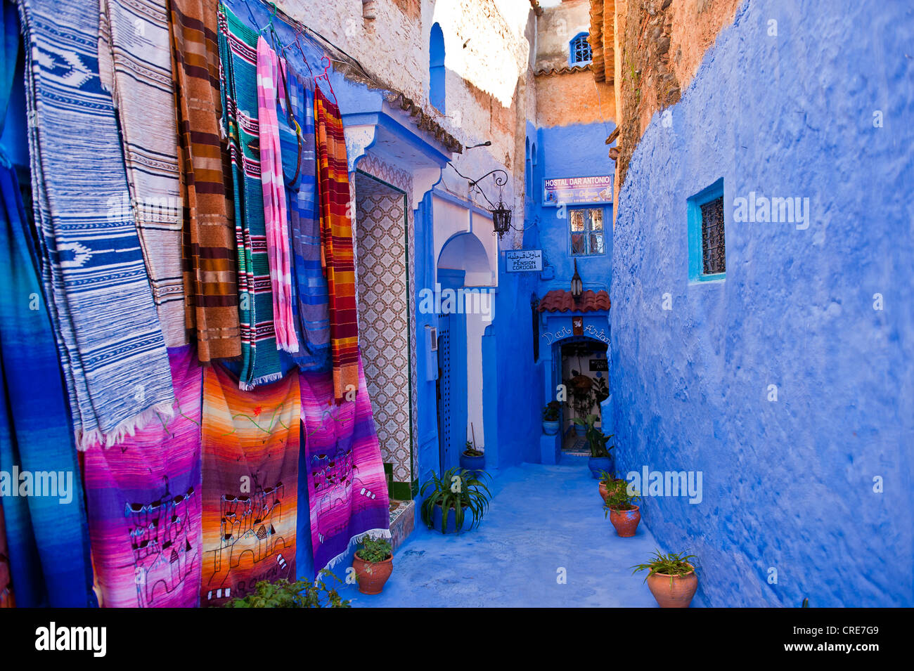 Narrow alleyway in Chefchaouen, walls and footpath are painted in blue, colourful blankets for sale hanging on the wall Stock Photo