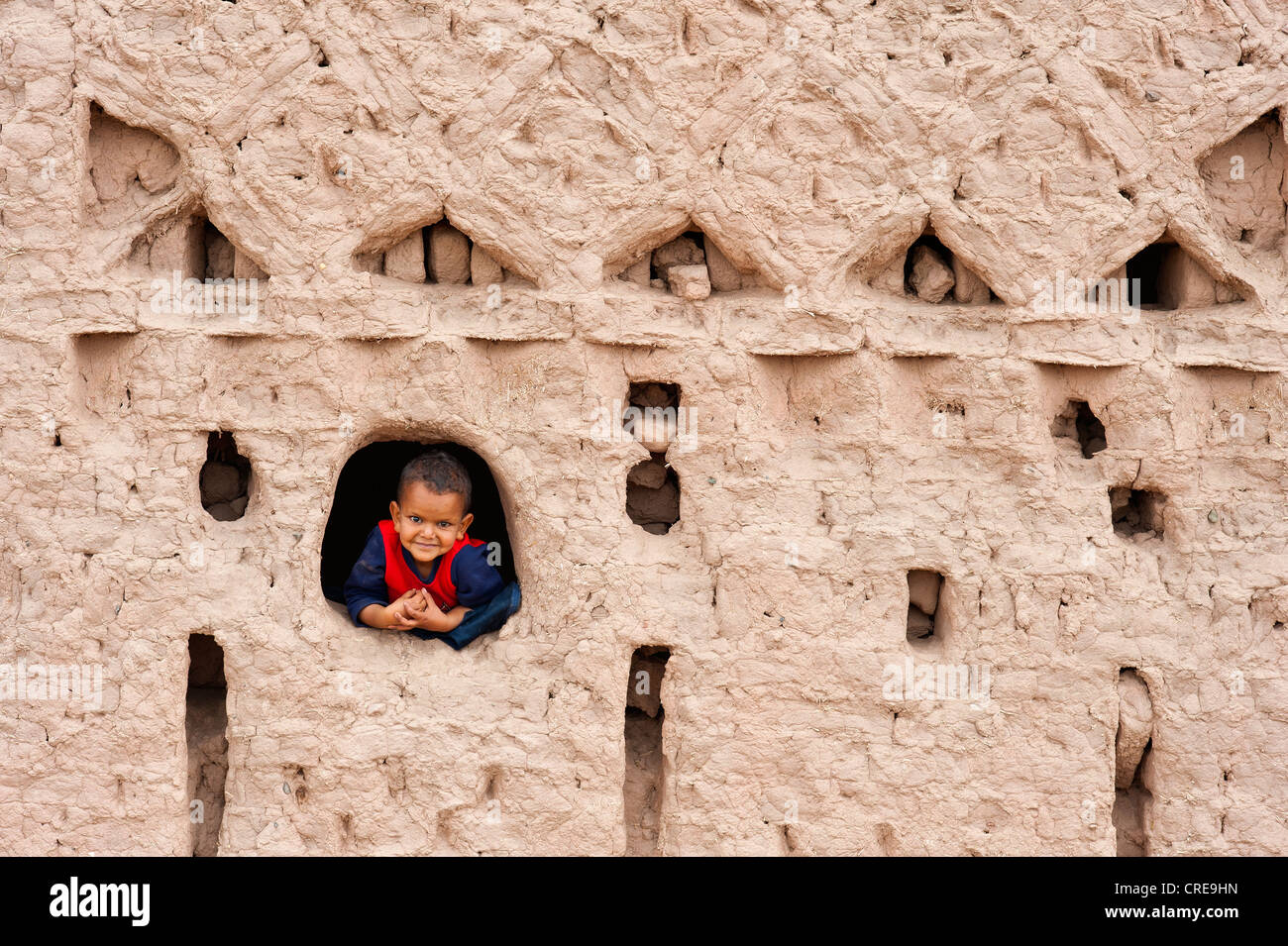 Young boy looking curiously out of a window of a kasbah, Thigremt, built of rammed earth, residential castle of the Berbers Stock Photo