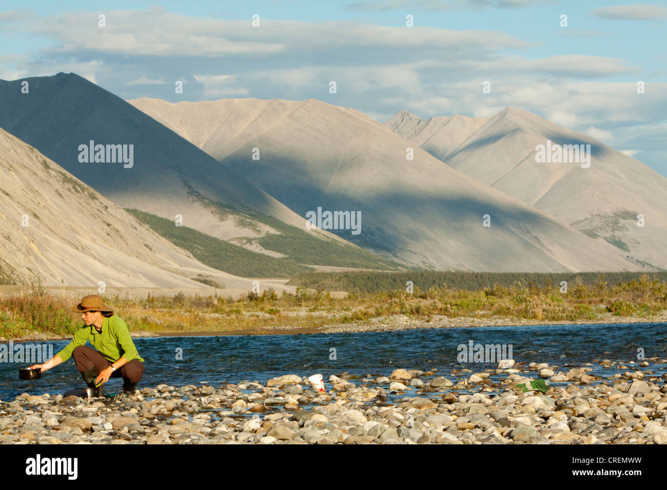 Young woman washing dishes in a river, camping, gravel bar, Mackenzie Mountains behind, Wind