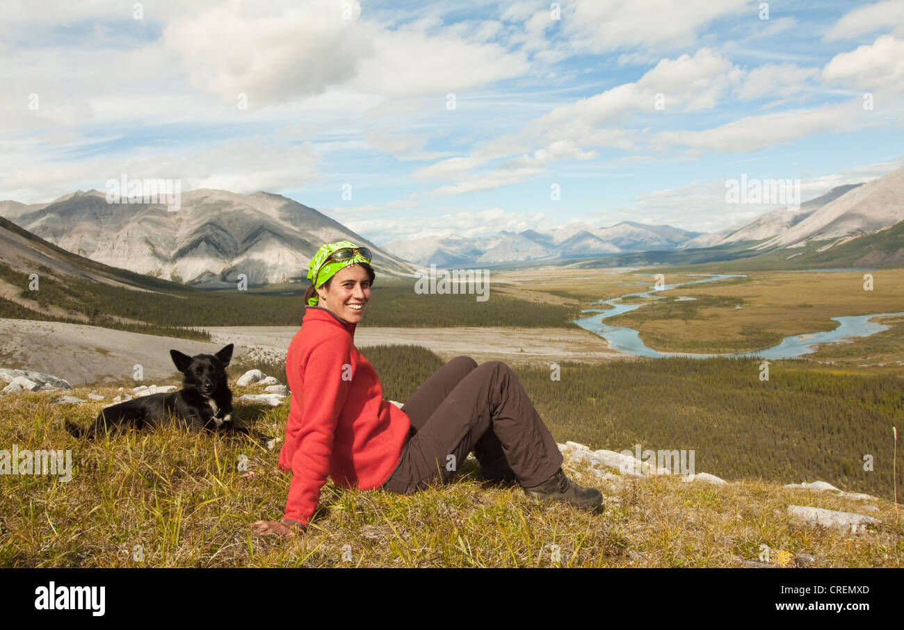 Young woman sitting, resting, enjoying the view, panorama, Alaskan Husky, sled dog beside her, Wind River valley and Mackenzie Stock Photo