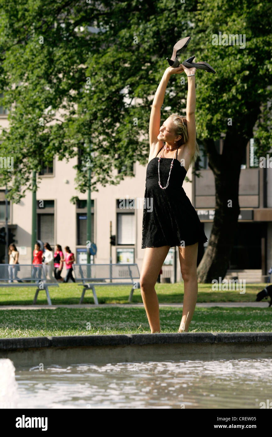 young blond woman wearing black summer dress standing near fountain, Germany Stock Photo