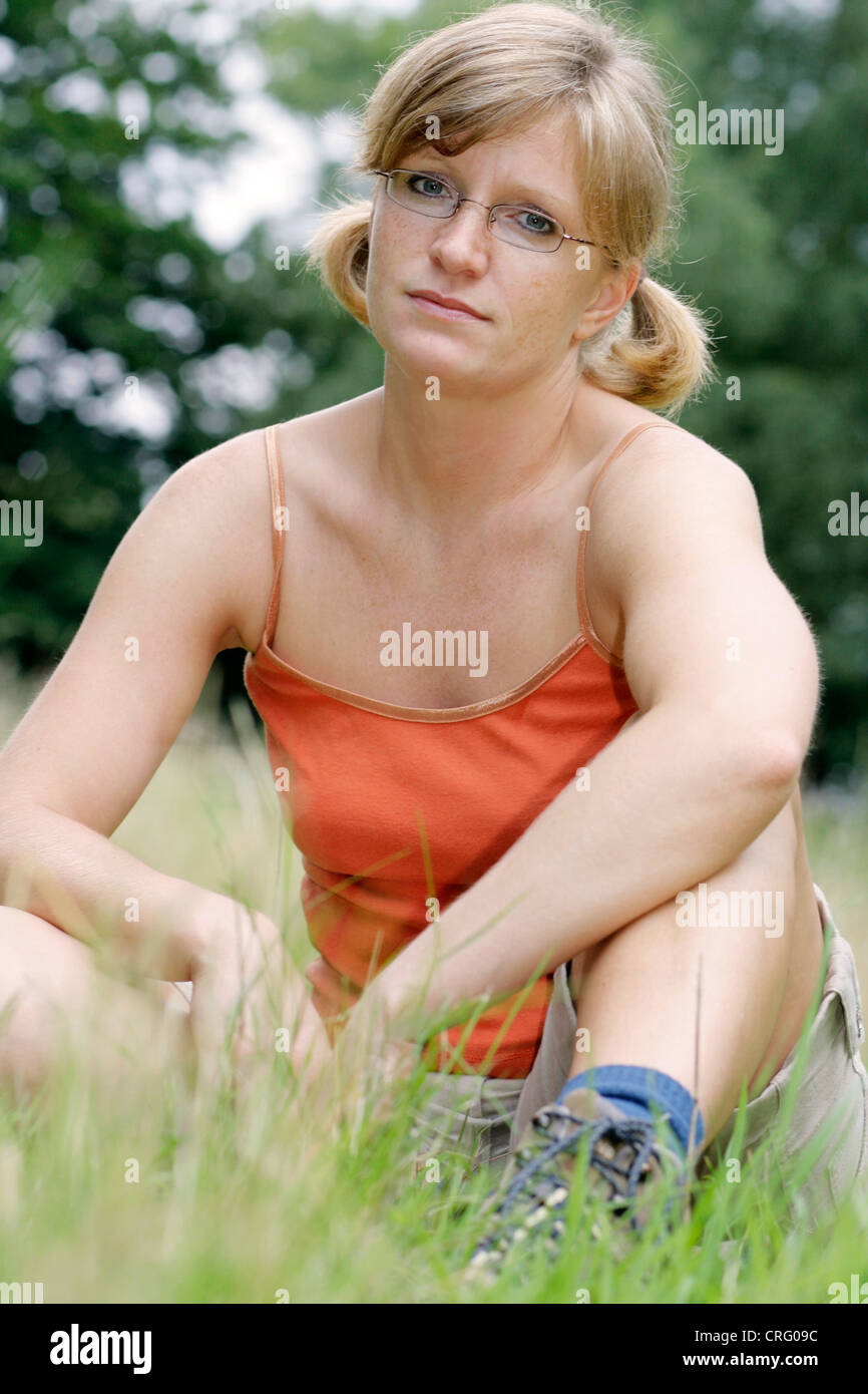 reflective young blond, sportive woman wearing glasses and casual wear, sitting in a meadow Stock Photo