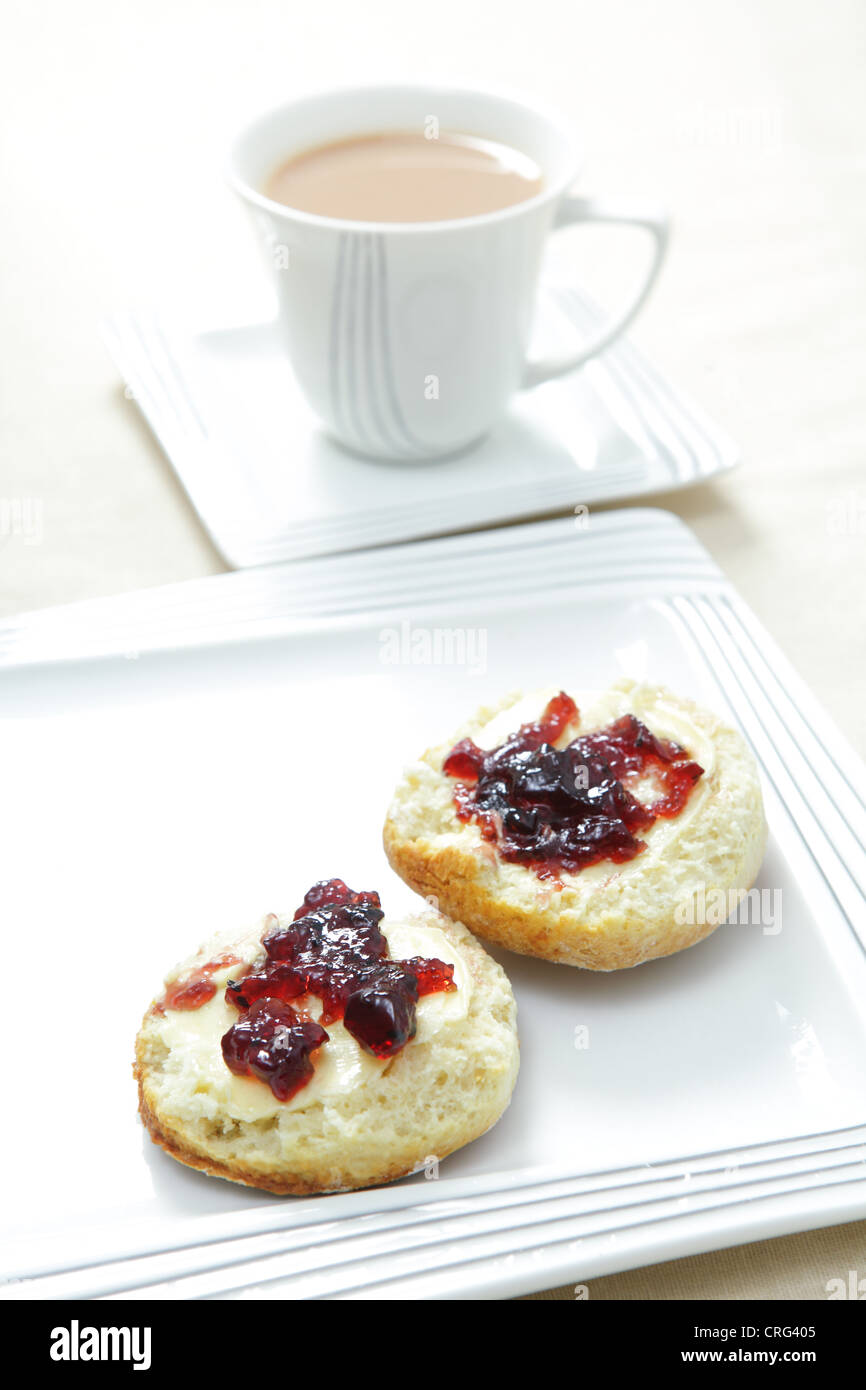 A traditional English afternoon tea of a scone topped with butter and blackcurrant jam and a cup of milky tea. Stock Photo