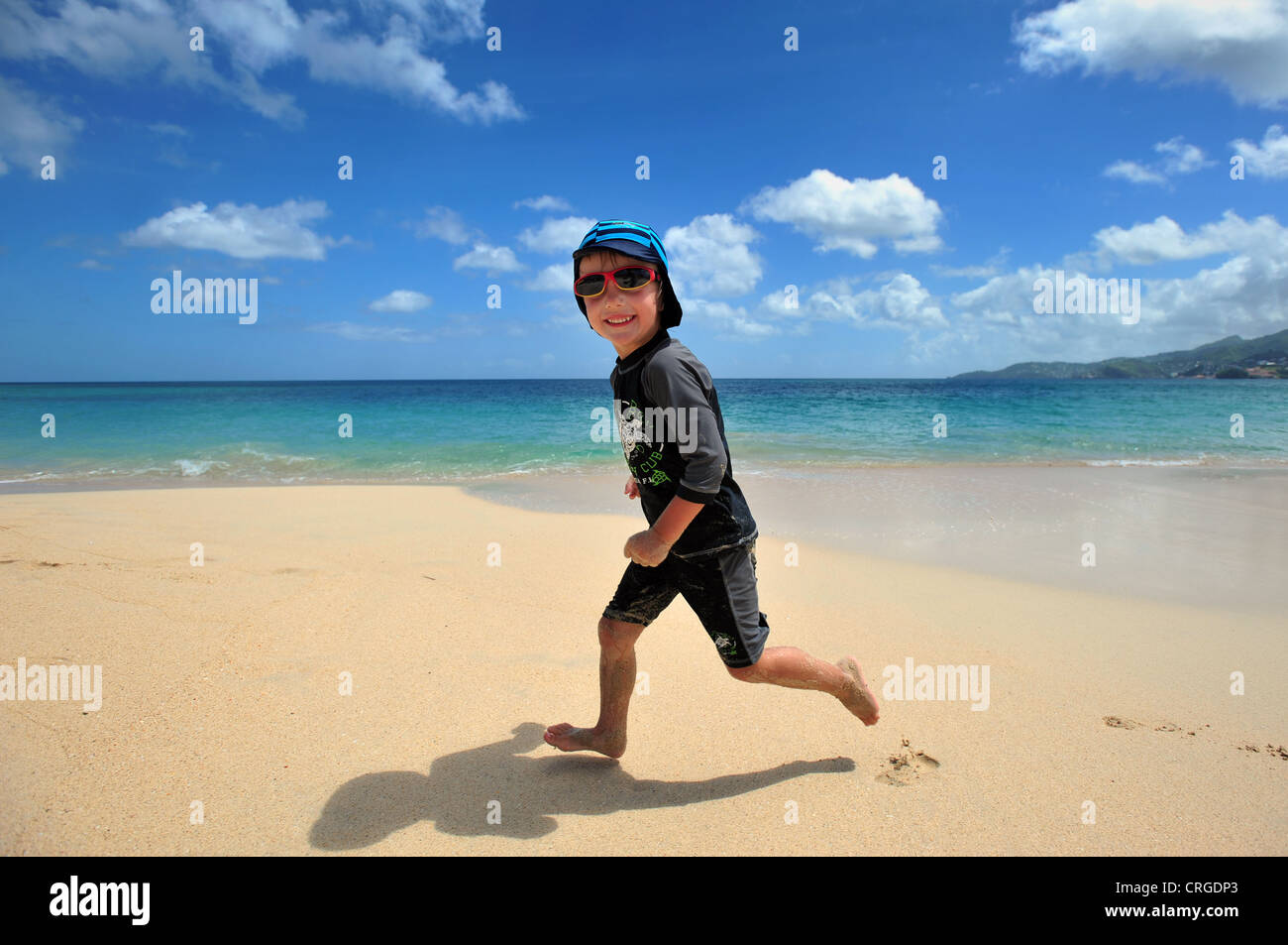A four year old boy wearing sun suit and sunglasses runs on the beach, Grenada, Caribbean, West Indies Stock Photo