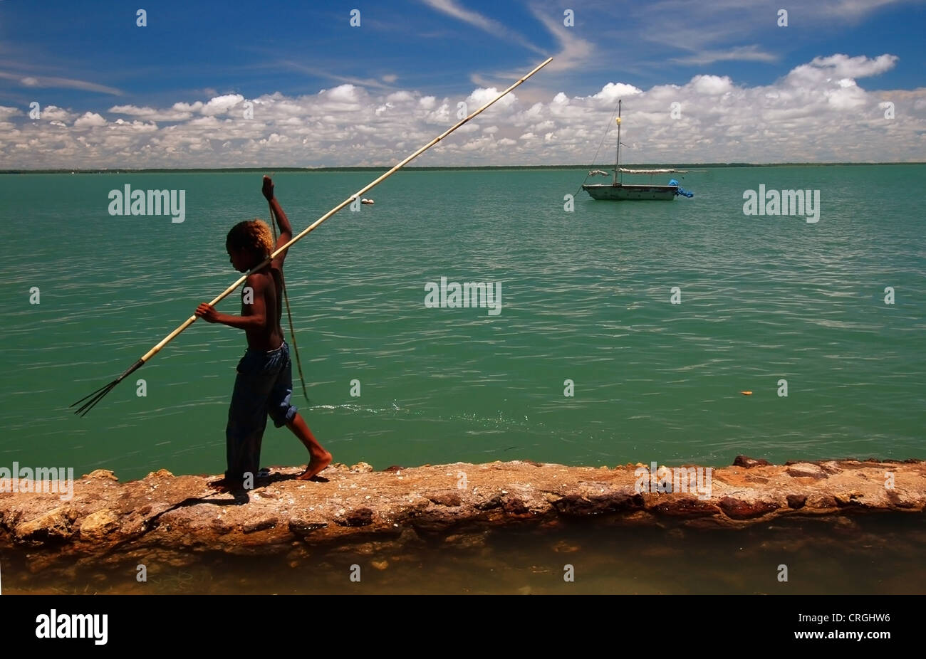 Boy with fish spear walks along ineffective seawall, Saibai Island, Torres Strait, Australia. Papua New Guinea on horizon Stock Photo