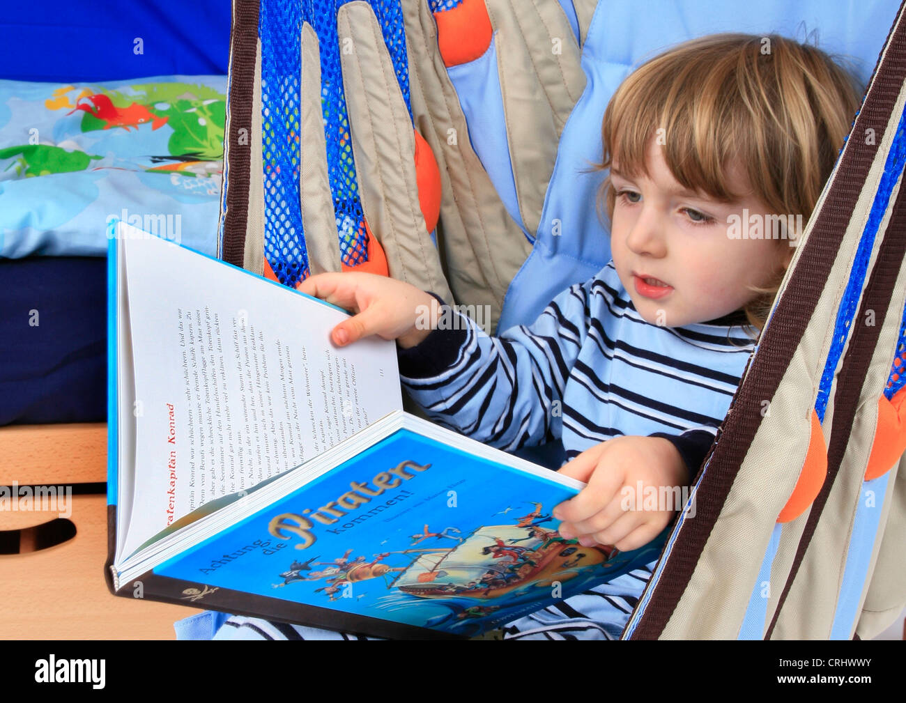 little boy in pyjama reading a children's book about pirates in a swing of a Billi-Bolli loft bed Stock Photo