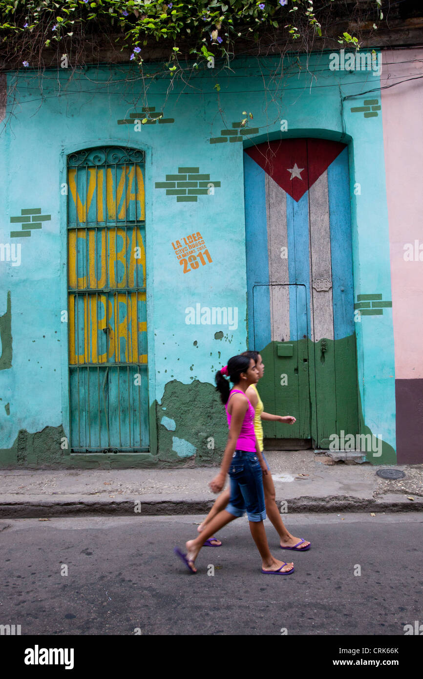 Two girls walking down the street in Old Havana Cuba, with a Cuban flag and viva Cuba libre painted on two doors in background. Stock Photo