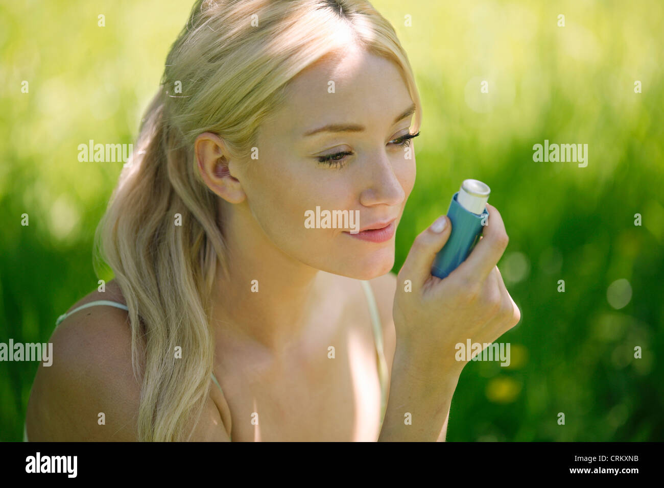 A young blond woman sitting in the grass using a asthma inhaler Stock Photo