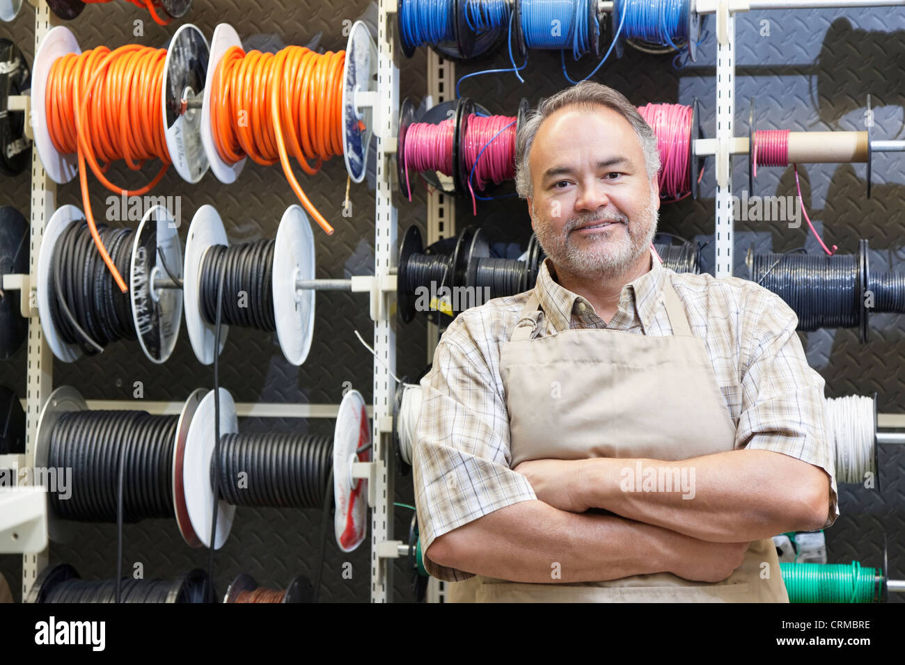 Portrait of a happy mature salesperson standing in front of electrical wire spool with arms crossed in hardware store Stock Photo