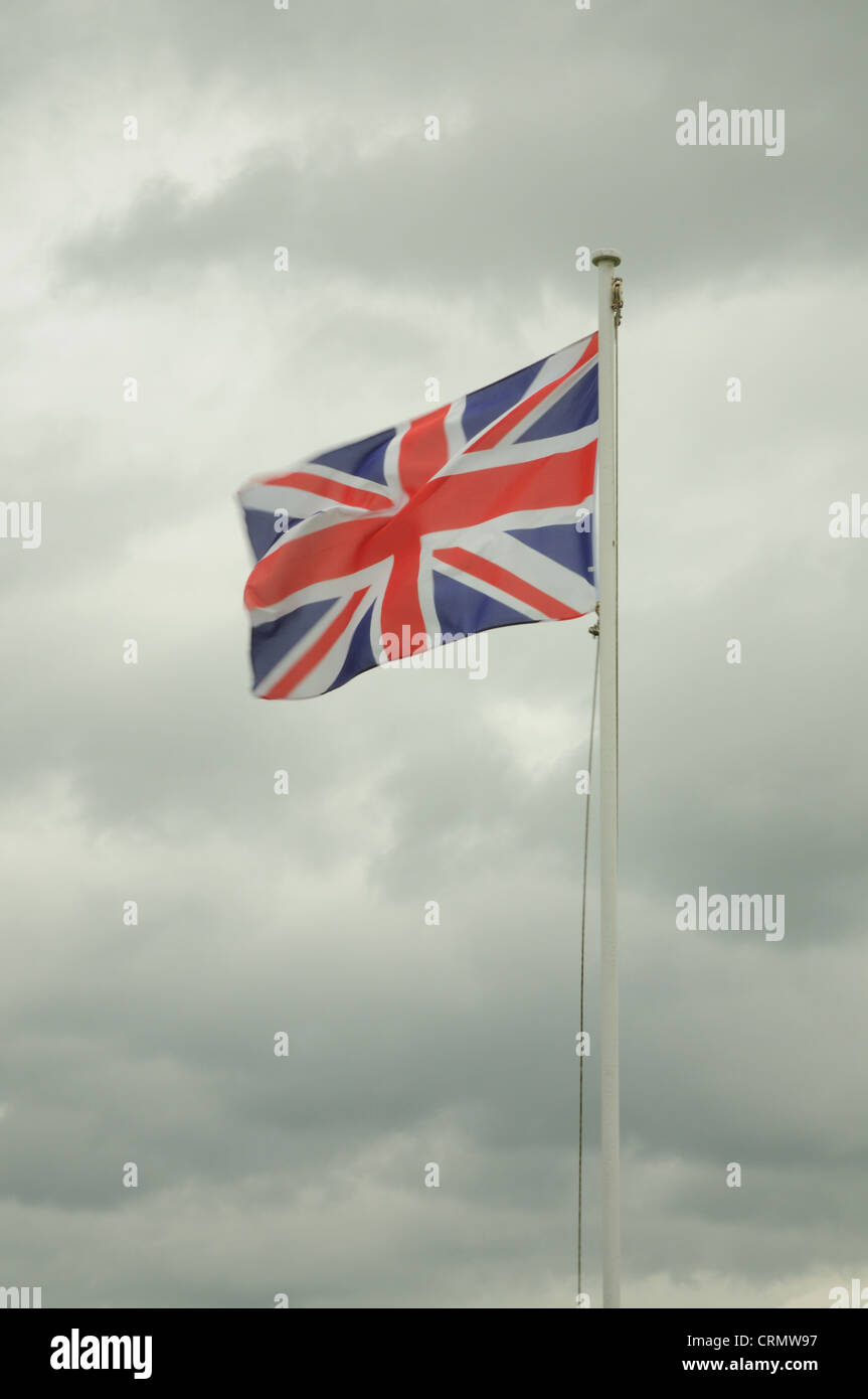 Union Jack Flag of the United Kingdom on a Flag Pole, blowing in the wind with a very cloudy and gloomy sky Stock Photo