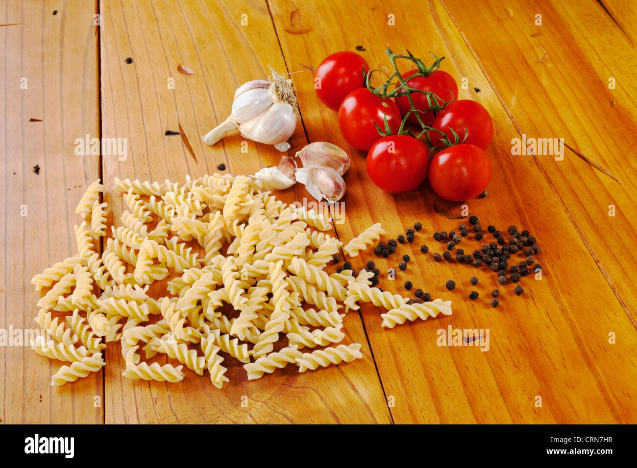Uncooked fusili pasta with truss tomatoes, garlic and black peppercorns, on an old wooden kitchen table. Stock Photo