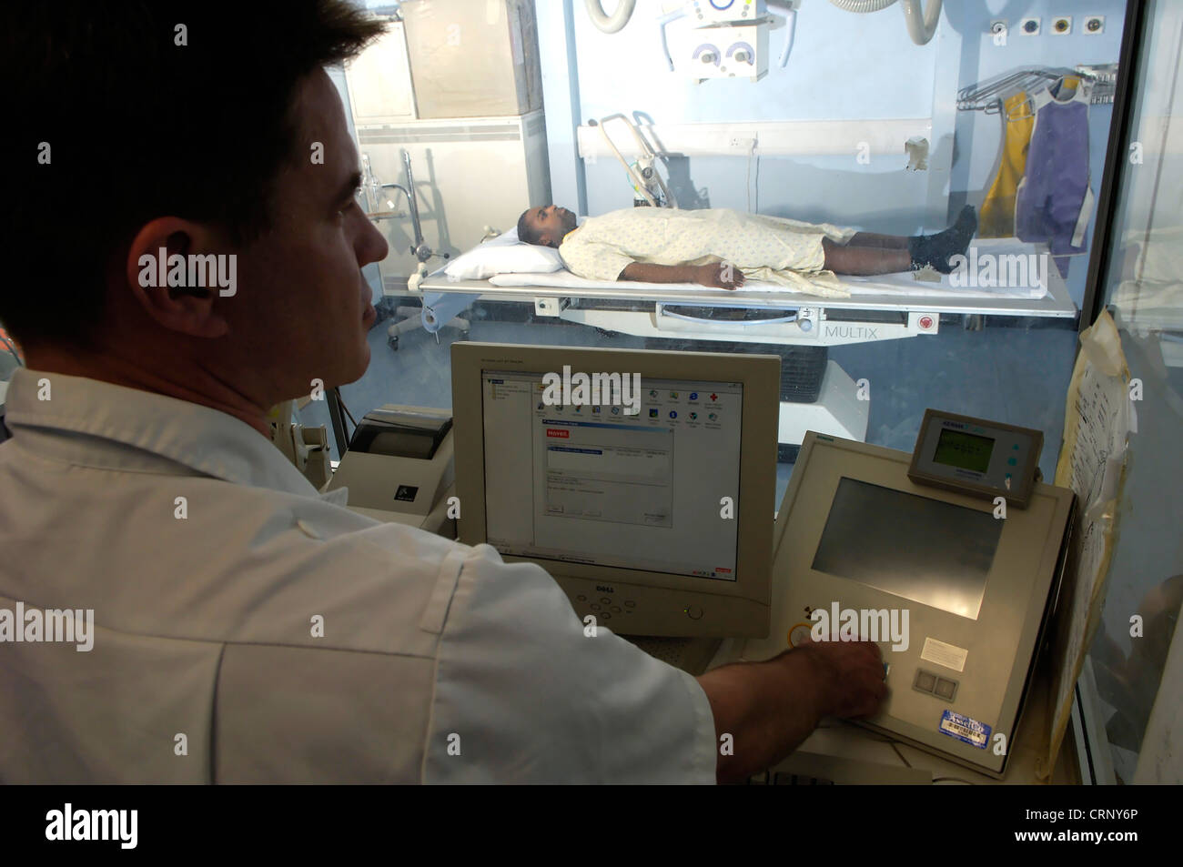 A radiologist sitting behind glass as a protection against radioactivity as he x-rays a patient lying on a bed. Stock Photo