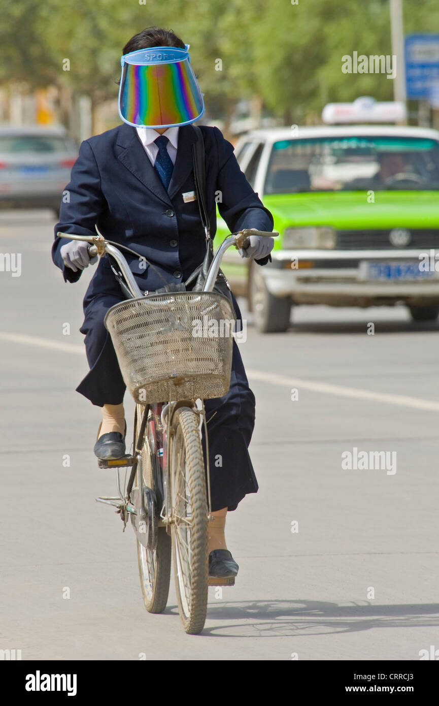 A Chinese woman travels along a road on her bicycle in Kashgar wearing a sun shield visor. Stock Photo