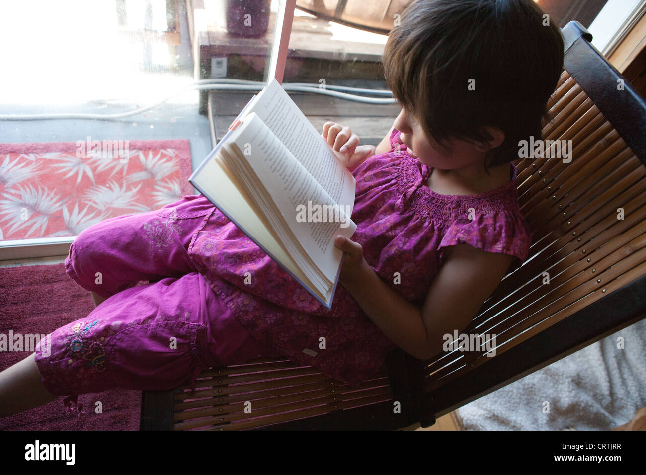 Seven year old girl reading an adult level book in a chair at home. Stock Photo