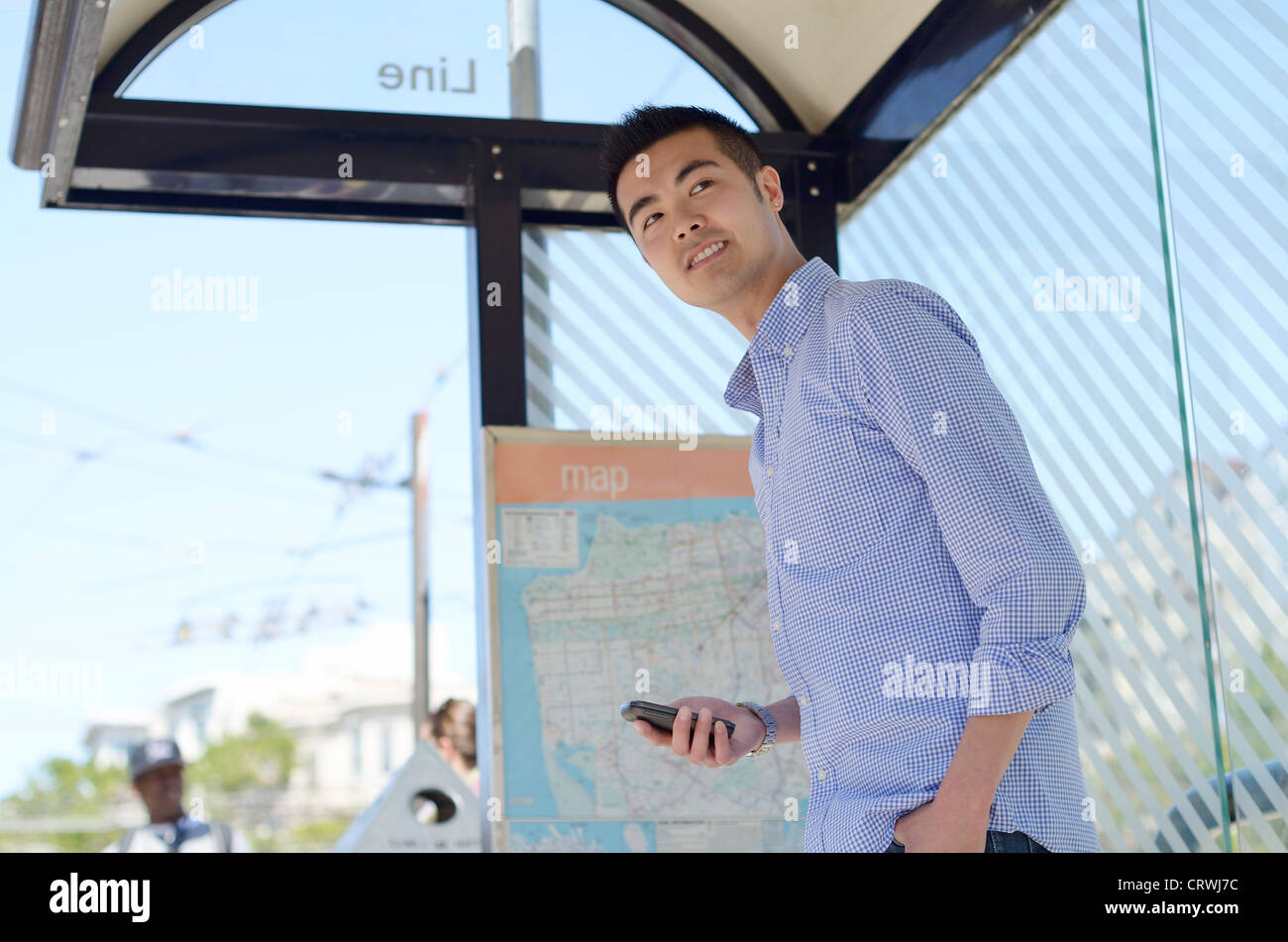 Young Asian man looks out from a bus stop with his phone in hand Stock Photo