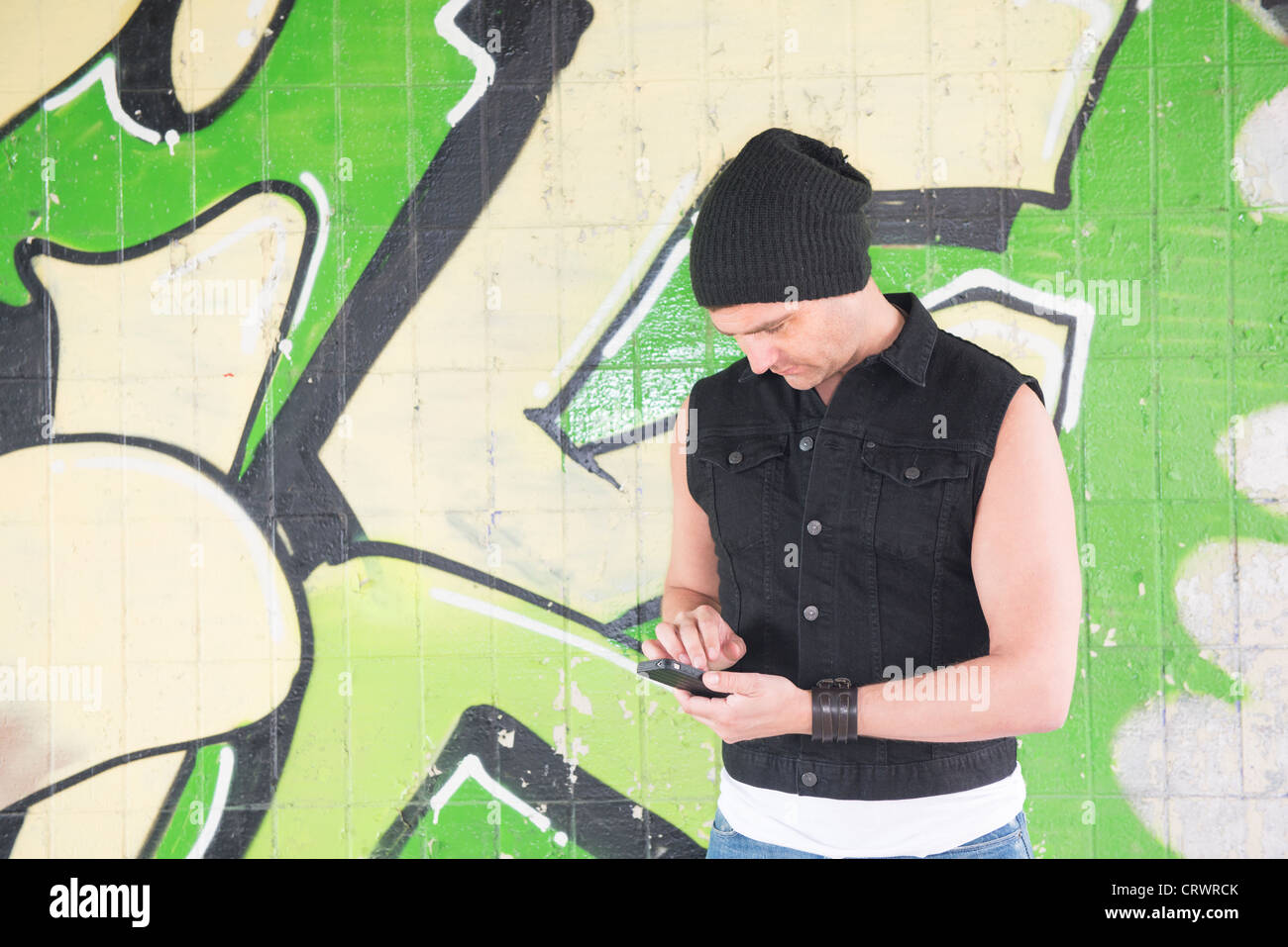 Man wearing a beanie and standing in front of graffitti wall is sending a message on his cellphone Stock Photo