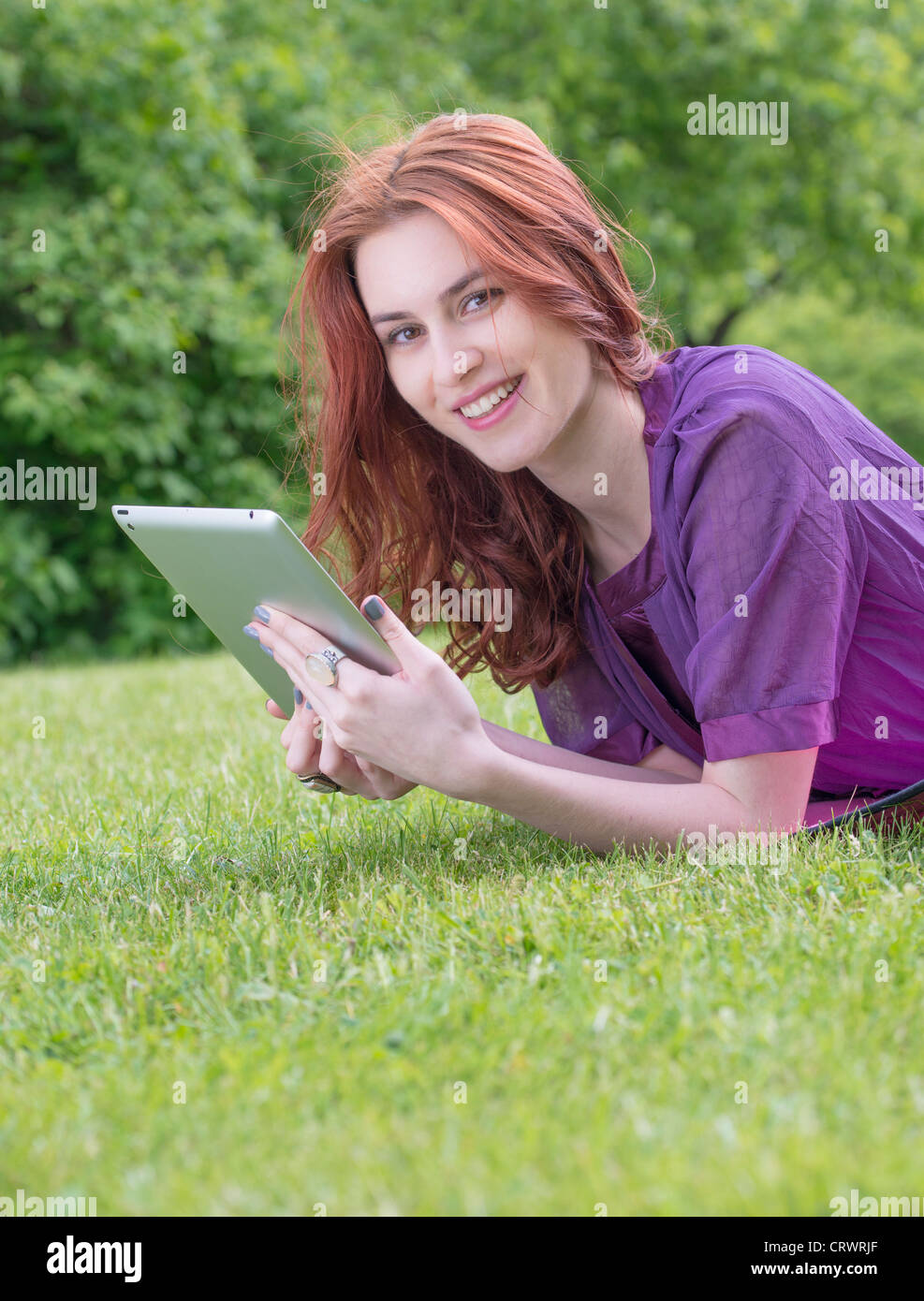 Happy young female adult with red hair lying on a lawn holding a tablet pc Stock Photo