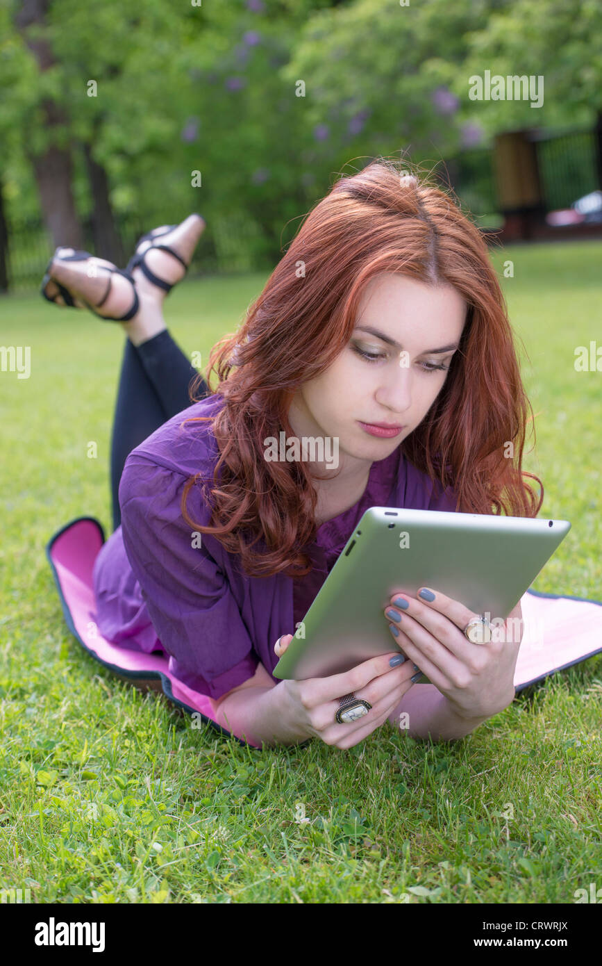 Young female adult with red hair lying on a lawn looking at a tablet pc Stock Photo