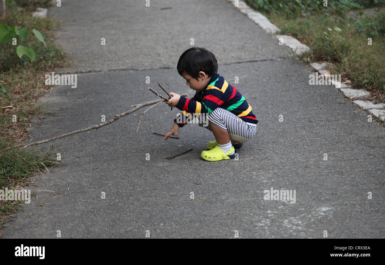 A little Chinese Boy i splaying with a wood stick in a walk park in Hong Kong. He wears a pull over with colorful stripes Stock Photo