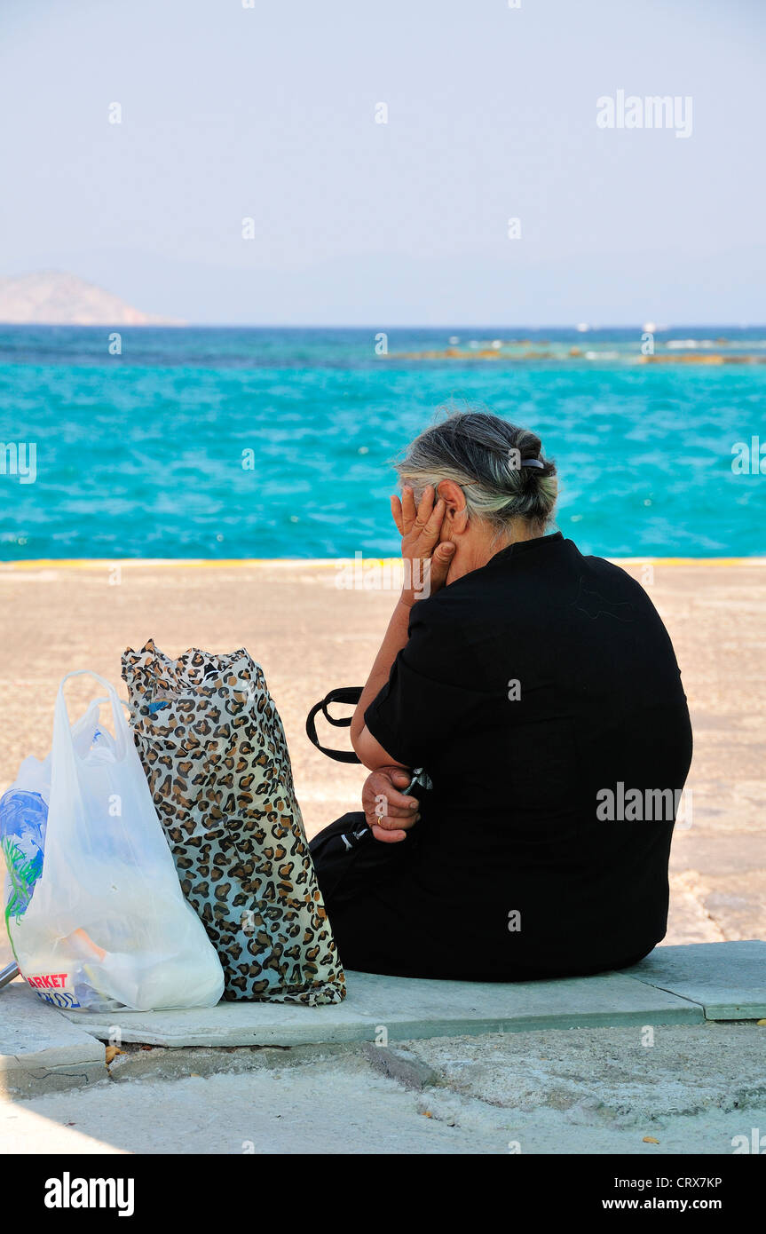 Grey haired elderly Greek lady dressed in black with head in hand sitting on the quay waiting for ferry at the main port on Aegina Island, Greece Stock Photo