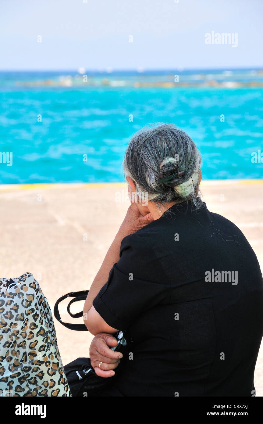 Rear view of old Greek lady dressed in black waiting for ferry sitting on the quay at the main port on Aegina Island, Greece Stock Photo