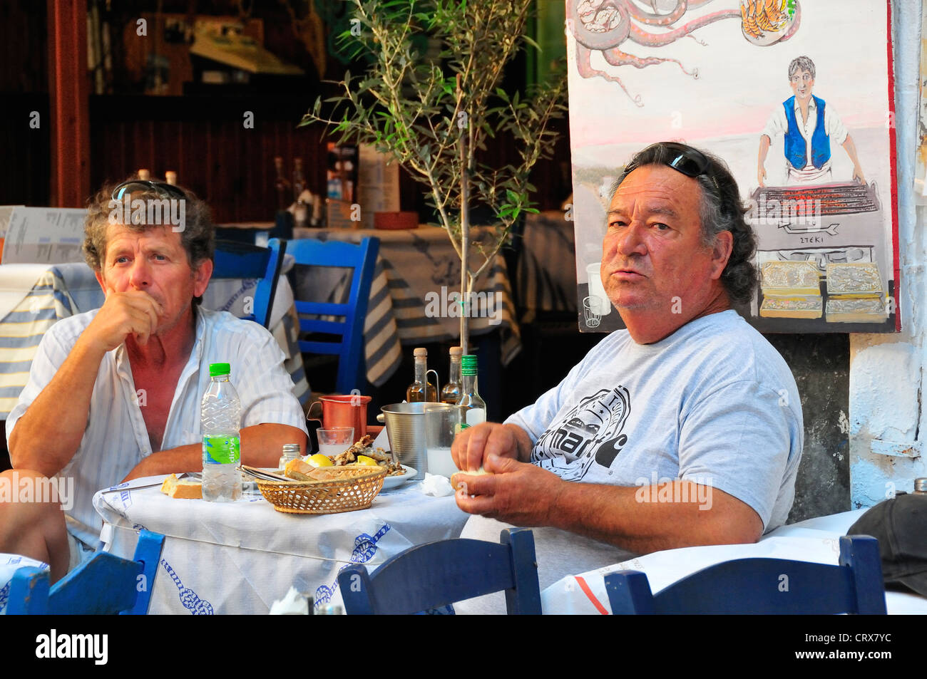 Two Greek men on the island of Aegina, Greece, sit in taverna with blue chairs in the main town enjoying a meal at lunch time Stock Photo