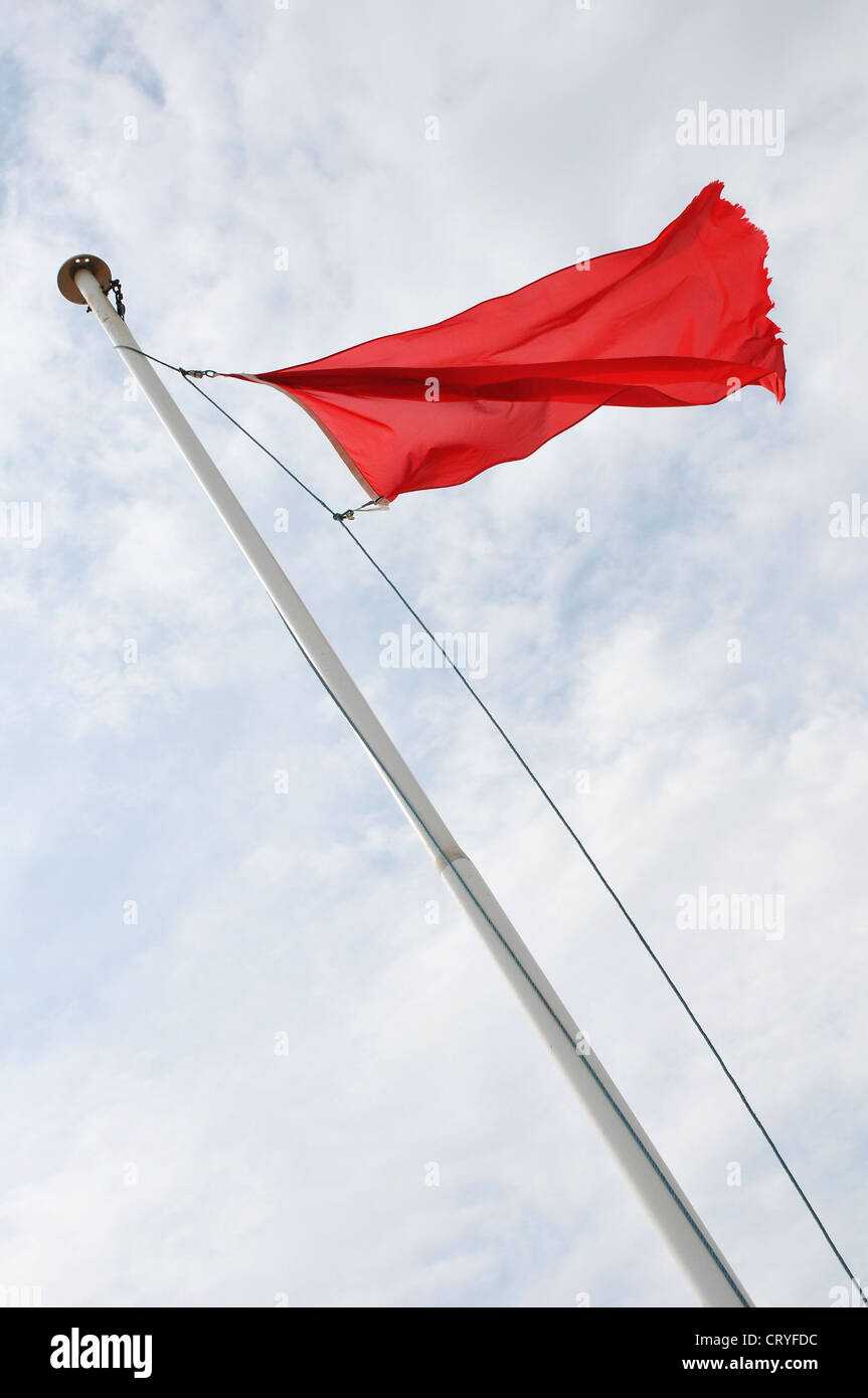 A red flag flying from flag pole seen from below, clouds and blue sky beyond Stock Photo
