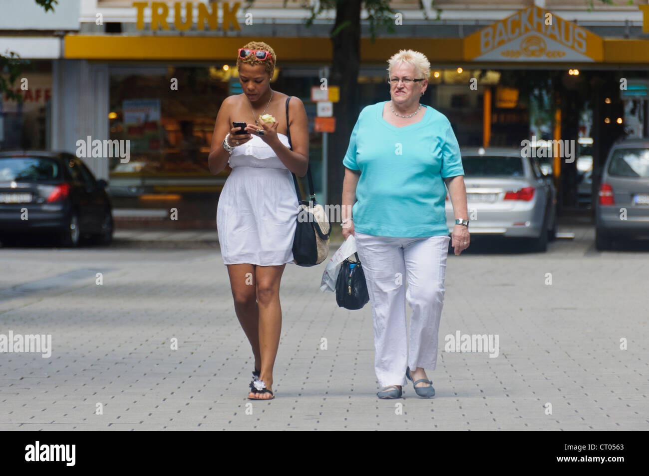 Elderly Caucasian Woman looks enviously at Brown Mixed Race Black Woman eating Apple use Smartphone - Heilbronn, Germany Stock Photo