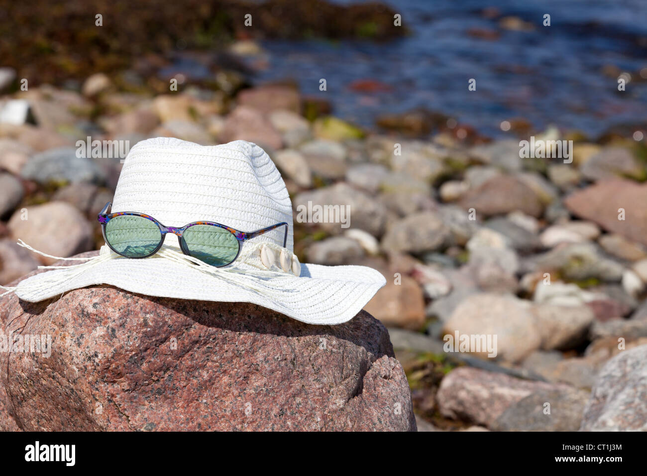 An white straw hat with sunglasses lies on a red big rock on the beach ...