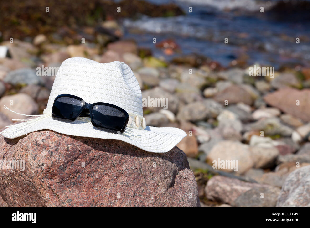 An white straw hat with sunglasses lies on a red big rock on the beach ...